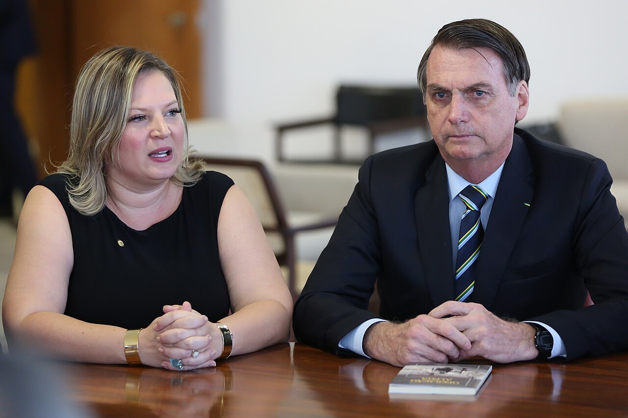 Joice Hasselmann, de vestido preto, fala sentada a uma mesa de madeira ao lado de Jair Bolsonaro, de terno escuro e gravata listrada, no Palácio do Planalto em março de 2019